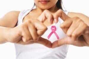 Woman in a white tank top forming a heart shape, wearing a pink ribbon for breast cancer awareness.
