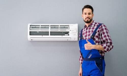 HVAC technician in blue overalls giving thumbs-up by wall-mounted air conditioning unit at Barker Heating & Cooling service.