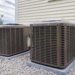 Two weathered air conditioning units installed on a stone surface against a building wall by Barker Heating & Cooling.