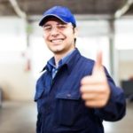 A smiling HVAC worker in a blue uniform and cap gives a thumbs-up in an industrial workshop setting.