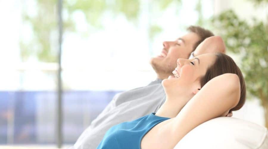 A man and woman reclining on a couch, smiling in a bright indoor space, promoting comfort and HVAC readiness.