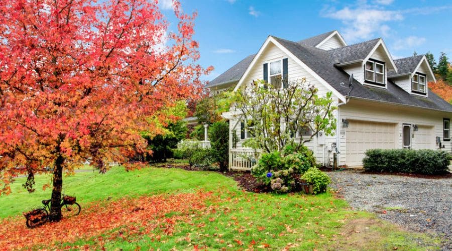 White two-story house with porch and vibrant autumn tree, showcasing Barker Heating & Cooling's focus on home comfort.
