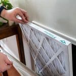 Person’s hands replacing a white air filter in a tidy home with wooden furniture, showcasing HVAC maintenance essentials.