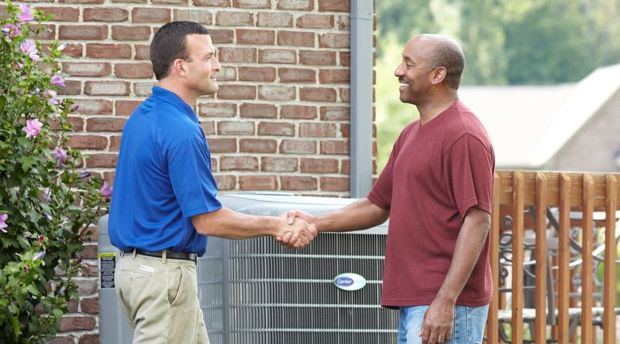 Service technician and homeowner shake hands beside an HVAC air conditioning unit in a residential setting.