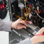 Technician repairing HVAC system with multimeter, inspecting wiring and machine components for safety issues.