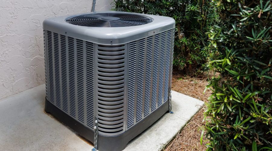 Gray air conditioning unit on a concrete slab, surrounded by green shrubbery, highlighting HVAC home comfort solutions.